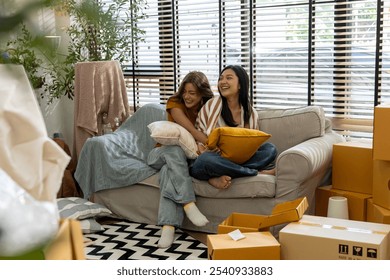 Two young asian women embrace and laugh together on a cozy couch, surrounded by moving boxes in a bright, well-lit living room. Happy Lesbian Couple Relaxing in Their New Home After Moving Day - Powered by Shutterstock