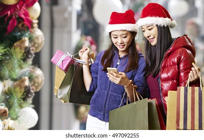 Two Young Asian Women Looking At Mobile Phone During Christmas Shopping