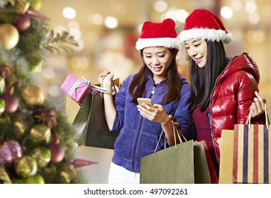 Two Young Asian Women Looking At Mobile Phone During Christmas Shopping