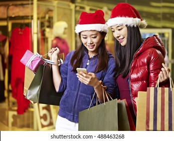 Two Young Asian Women Wearing Christmas Hat Carrying Shopping Bags Looking At Cellphone