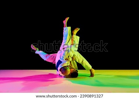 Two young, athletic professional karate men fighting, performing technical skill in neon light isolated black background. Concept of martial art, combat sport, health, strength, energy. Copy space