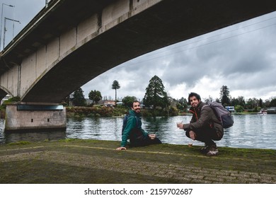 Two Young, Bearded, Happy And Handsome Brothers With Jacket And Backpack Eating Ceviche (fresh Raw Fish With Citrus Juice And Chili Peppers) And Panoramic View Of River, Bridge And Cloudy Sky,Valdivia