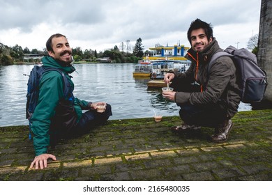 Two Young, Bearded, Happy And Handsome Brothers With Jacket And Backpack Eating Ceviche (fresh Raw Fish With Citrus Juice And Chili Peppers) And Panoramic View Of River, Boats And Cloudy Sky, Valdivia