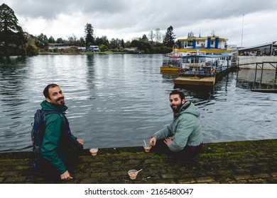 Two Young, Bearded, Happy And Handsome Brothers With Jacket And Backpack Eating Ceviche (fresh Raw Fish With Citrus Juice And Chili Peppers) And Panoramic View Of River, Boats And Cloudy Sky, Valdivia