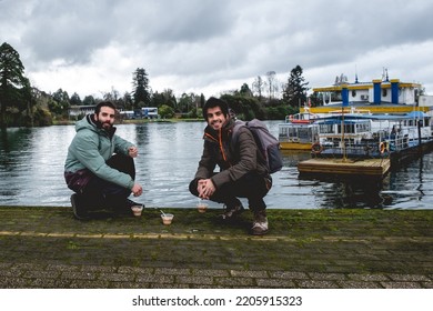 Two Young, Bearded, Happy And Handsome Brothers With Jacket And Backpack Eating Ceviche (fresh Raw Fish With Citrus Juice And Chili Peppers) And Panoramic View Of River, Boats And Cloudy Sky, Valdivia