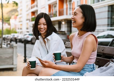 Two young beautiful asian laughing girls with coffee with closed eyes sitting on a bench in the street - Powered by Shutterstock