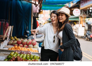 Two Young Beautiful Asian Women Friends Travel In Taiwan Taipei Local Market Shopping In Fruit Vendor. Lady Point Finger To Fresh Vegetables And Showing Sisters. Female Discussing What To Buy.