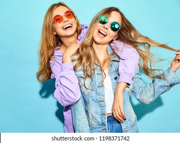 Two young beautiful blond smiling hipster girls in trendy summer clothes. Sexy carefree women posing near blue wall in sunglasses. Positive models having fun