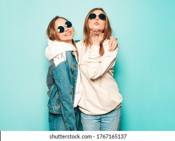 Two young beautiful brunette smiling hipster girls in trendy summer hoodie and jeans jacket clothes. Sexy carefree women posing near blue wall. Trendy and positive models give air kiss in sunglasses