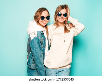Two young beautiful brunette smiling hipster girls in trendy summer hoodie and jeans jacket clothes. Sexy carefree women posing near blue wall. Trendy and positive models having fun in sunglasses