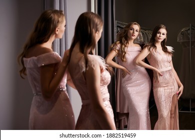 Two Young Beautiful Girls Wearing Full-length Pink Violet Or Purple Chiffon Mermaid Dresses Or Prom Ball Gowns Decorated With Sparkles And Sequins. Models In Front Of Mirror In A Fitting Room At Dress