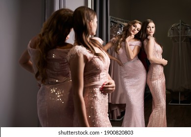 Two Young Beautiful Girls Wearing Full-length Pink Violet Or Purple Chiffon Mermaid Dresses Or Prom Ball Gowns Decorated With Sparkles And Sequins. Models In Front Of Mirror In A Fitting Room At Dress