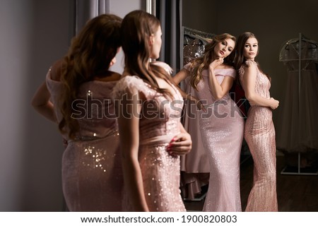 Two young beautiful girls wearing full-length pink violet or purple chiffon mermaid dresses or prom ball gowns decorated with sparkles and sequins. Models in front of mirror in a fitting room at dress