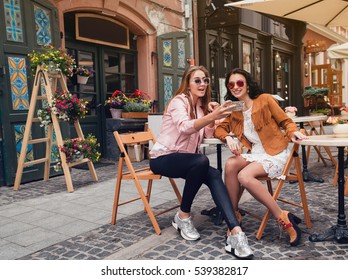two young beautiful hipster women sitting at cafe, stylish trendy outfit, europe vacation, street style, happy, having fun, smiling, sunglasses, looking at smartphone, taking selfie photo, flirty