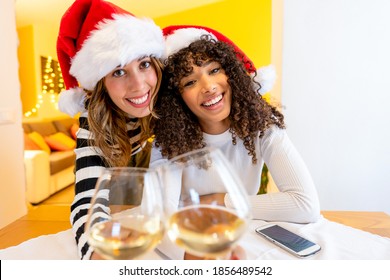 Two young beautiful mixed race women wearing Santa hat looking at the camera holding white wine glasses - Female lgbt couple smiling toasting with champagne in video call for Christmas and New Year - Powered by Shutterstock