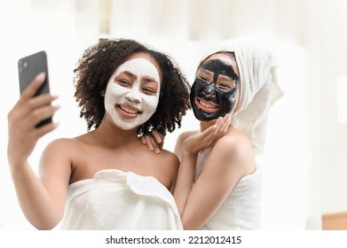 Two Young Beautiful Multi Ethnic Asian And African Woman LGBT Couple Or Friend Using Smartphone Camera Taking Selfie Photo Together After Applied Beauty Skin Care Moisturizer Cream Face Mask Happily.