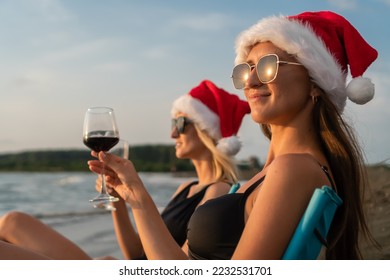 Two young beautiful slender women in swimsuits, sunglasses, Santa Claus hats are sitting on folding chairs on the sandy beach with glasses of red wine at sunset. Focus on the brown-haired woman - Powered by Shutterstock