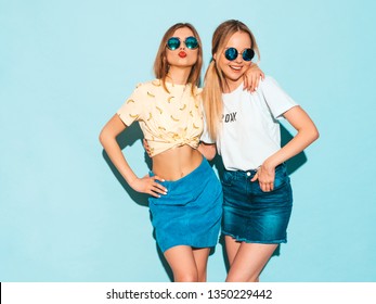 Two young beautiful smiling blond hipster girls in trendy summer colorful T-shirt clothes. Sexy carefree women posing near blue wall in round sunglasses. Positive models having fun