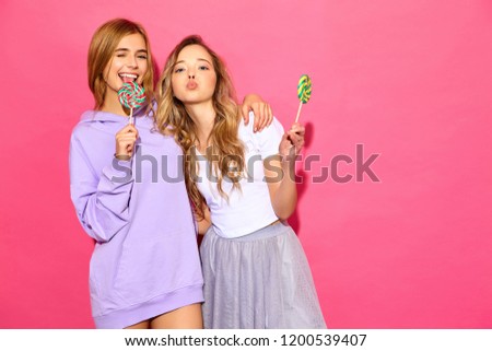 Two young beautiful smiling blond hipster girls in trendy summer clothes. Carefree hot women posing near pink wall. Positive funny models with lollipop, winking