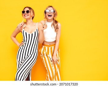 Two Young Beautiful Smiling Brunette Hipster Female In Trendy Summer Clothes. Sexy Carefree Women Posing Near Yellow Wall In Studio. Positive Models Having Fun. Cheerful And Happy. In Sunglasses