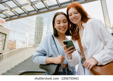 Two young beautiful smiling girls looking on the phone together, while standing outdoors - Shutterstock ID 2206733461