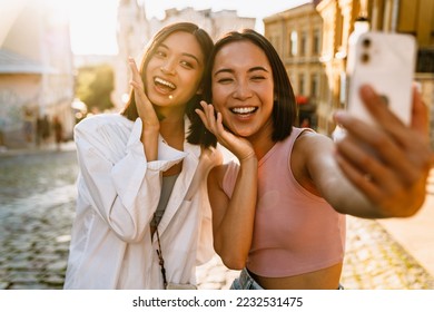 Two young beautiful smiling happy asian girls taking selfie on phone with raised hands near their cheeks , while standing on a sunny street - Powered by Shutterstock