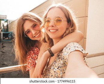 Two Young Beautiful Smiling Hipster Girls In Trendy Summer Sundress.Sexy Carefree Women Posing On The Street Background In Sunglasses. They Taking Selfie Self Portrait Photos On Smartphone At Sunset