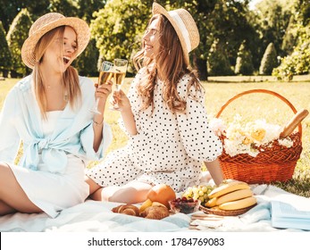 Two Young Beautiful Smiling Hipster Girls In Trendy Summer Sundress And Hats.Carefree Women Making Picnic Outside.Positive Models Sitting On Plaid On Grass,drinking Champagne, Eating Fruits And Cheese