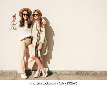 Two young beautiful smiling hipster female in trendy white sweater and coat. Sexy carefree women posing near wall on the street background in hat. Positive models having fun outdoors