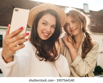 Two Young Beautiful Smiling Hipster Female In Trendy White Sweater And Coat. Sexy Carefree Women Posing On Street Background In Hat. Positive Models Having Fun Outdoors. Taking Photo Selfie