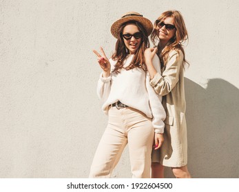 Two young beautiful smiling hipster female in trendy white sweater and coat. Sexy carefree women posing on  the street background in hat. Positive models having fun in sunglasses