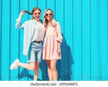 Two young beautiful smiling hipster female in trendy summer clothes and dress.Sexy carefree women posing in the street near blue wall.Positive pure models having fun at sunset, hugging and going crazy