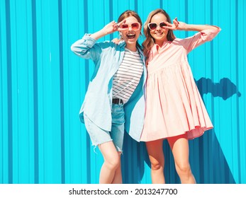 Two young beautiful smiling hipster female in trendy summer clothes and dress.Sexy carefree women posing in the street near blue wall.Positive pure models having fun at sunset, hugging.Show peace sign
