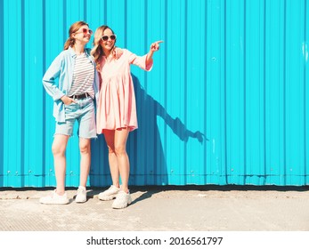 Two young beautiful smiling hipster female in trendy summer clothes and dress.Sexy carefree women posing in the street near blue wall.Positive pure models having fun at sunset, hugging