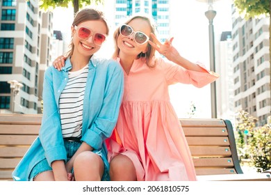 Two young beautiful smiling hipster female in trendy summer clothes and dress.Sexy carefree women posing in the street. Positive pure models having fun at sunset, sitting on a bench. Shows peace sign