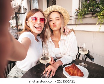 Two young beautiful smiling hipster female in trendy summer clothes.Carefree women posing at veranda cafe in the street.Positive models drinking white wine.Eating croissant.Taking Pov selfie photos - Powered by Shutterstock