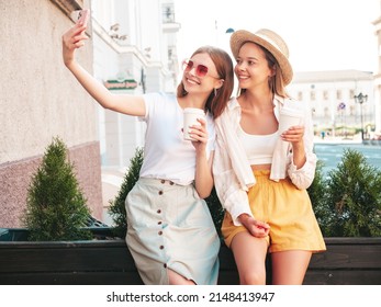 Two young beautiful smiling hipster female in trendy summer clothes.Sexy carefree women posing in the street. Positive pure models having fun at sunset. They drinking coffee or tea in plastic cup