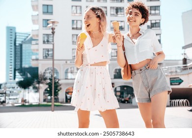 Two young beautiful smiling hipster female in trendy summer clothes. Carefree women posing on street background. Positive models eating tasty ice cream in waffles cone in sunny day, cheerful and happy - Powered by Shutterstock