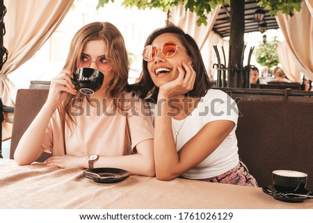 Two young beautiful smiling hipster girls in trendy summer clothes.Carefree women chatting in veranda terrace cafe and drinking coffee.Positive models having fun and communicating