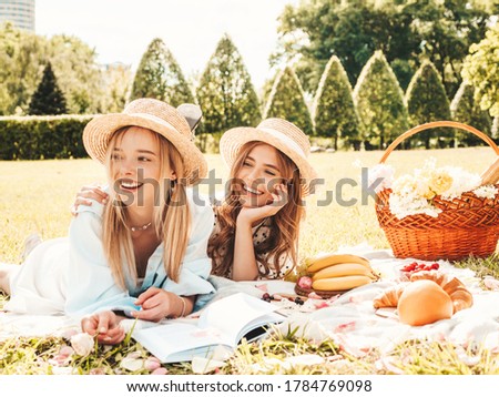 Two young beautiful smiling hipster girls in trendy summer sundress and hats.Carefree women making picnic outside.Positive models sitting on plaid on grass, reading book, eating fruits and cheese