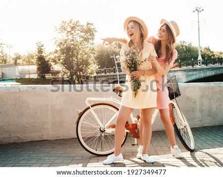 Two young beautiful smiling hipster woman in trendy summer sundress. Carefree women riding retro bicycle. Positive models having fun on bike posing at 
embankment in hats. Best friends outdoors