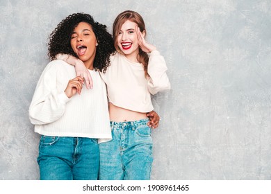 Two young beautiful smiling international hipster female in trendy summer jeans clothes. Sexy carefree women posing near gray wall in studio. Positive models having fun. Concept of friendship 