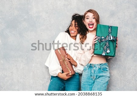 Two young beautiful smiling international hipster female in trendy clothes.Sexy carefree women posing near gray wall.Positive models hugging and giving each other gift boxes.Christmas, x-mas, concept