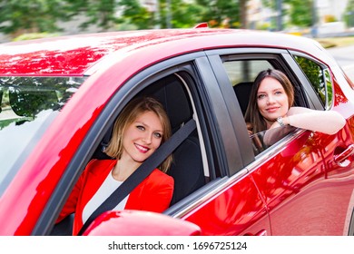 Two Young Beautiful Smiling Women Wearing Pink Sweater, Red Jacket And White T-shirt Driving Modern Car And Looking Into Camera Near Green Trees On A Summer Sunny Day. Travel And Friendship Concept