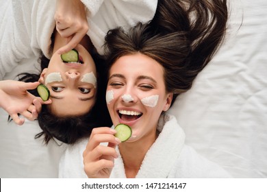 Two young beautiful women with dark hair in white bathrobes lying in bed with cosmetic mask on faces happily eating slices of cucumber together in modern cozy hotel  - Powered by Shutterstock