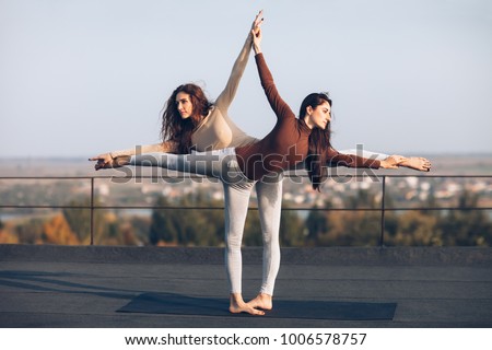 Two young beautiful women doing yoga asana virabhadrasana helping each other on the roof outdoor. partner yoga. Balance, concentration, equilibrium concept