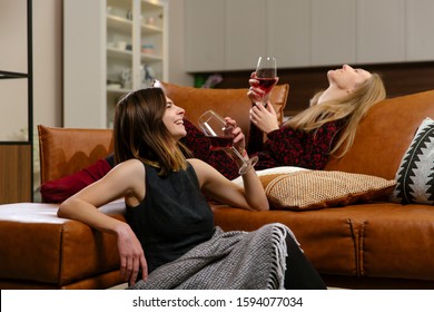 Two young beautiful women are drinking red wine, laughing, sitting on brown leather sofa with warm blanket, cushions, cozy atmosphere, wearing warm dresses and socks - Powered by Shutterstock