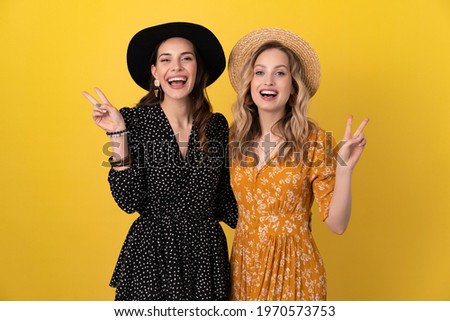 two young beautiful women friends together isolated on yellow background in black and yellow dress and hat stylish boho trend, spring summer fashion style accessories, smiling happy mood having fun