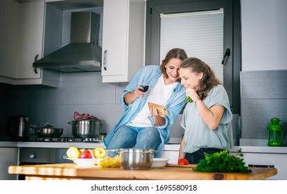 Two Young Beautiful Women Girlfriends Lesbian Couple Cook At Home In The Kitchen, Look At The Smartphone And Laugh Merrily, Home Activity, Cooking, Recipes, Stay At Home