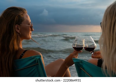 Two Young Beautiful Women In Swimsuits, Sunglasses Sitting On Folding Chairs On The Beach Clinking Glasses With Red Wine At Sunset, Photo From Behind